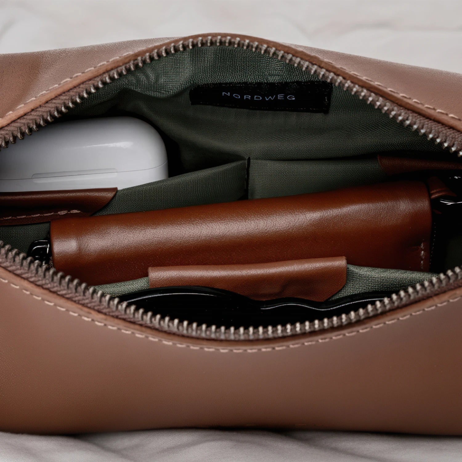 Open main compartment of the Nordweg Pulse Leather Sling Bag in caramel showing interior organization with an earbud case, pen, and accessories