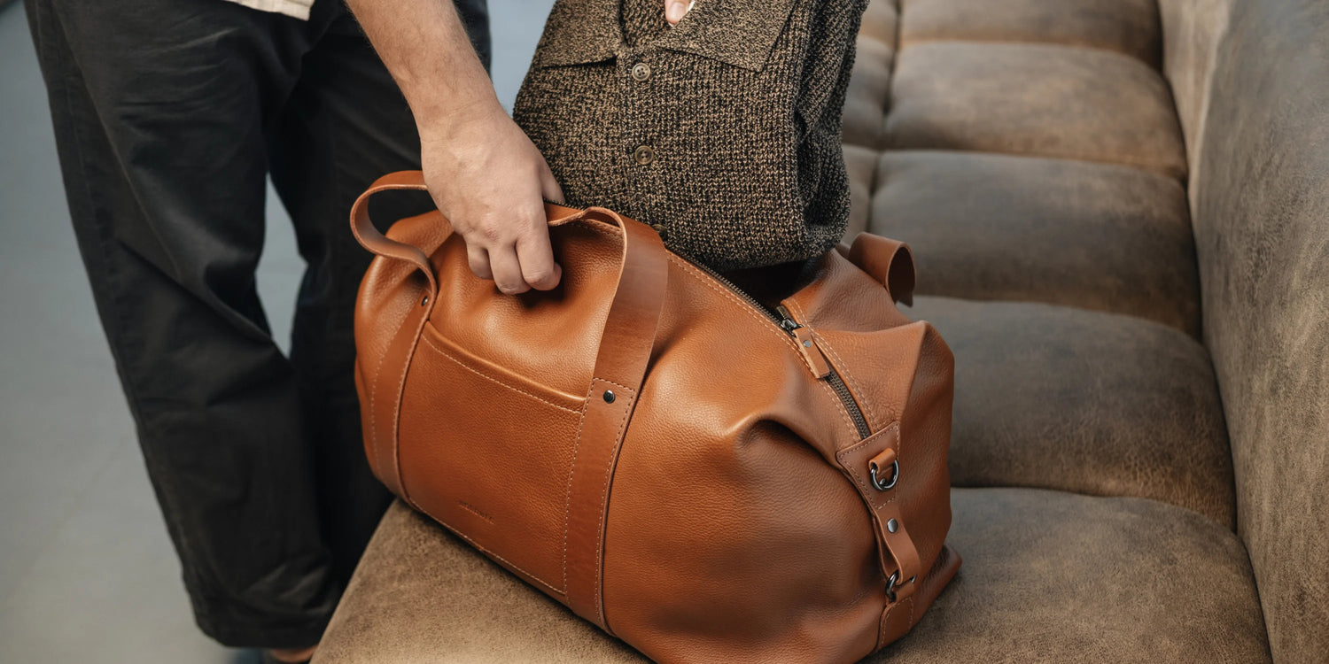 Person holding the Nordweg Legacy Leather Duffel Bag in cognac by its top handle, resting on a sofa