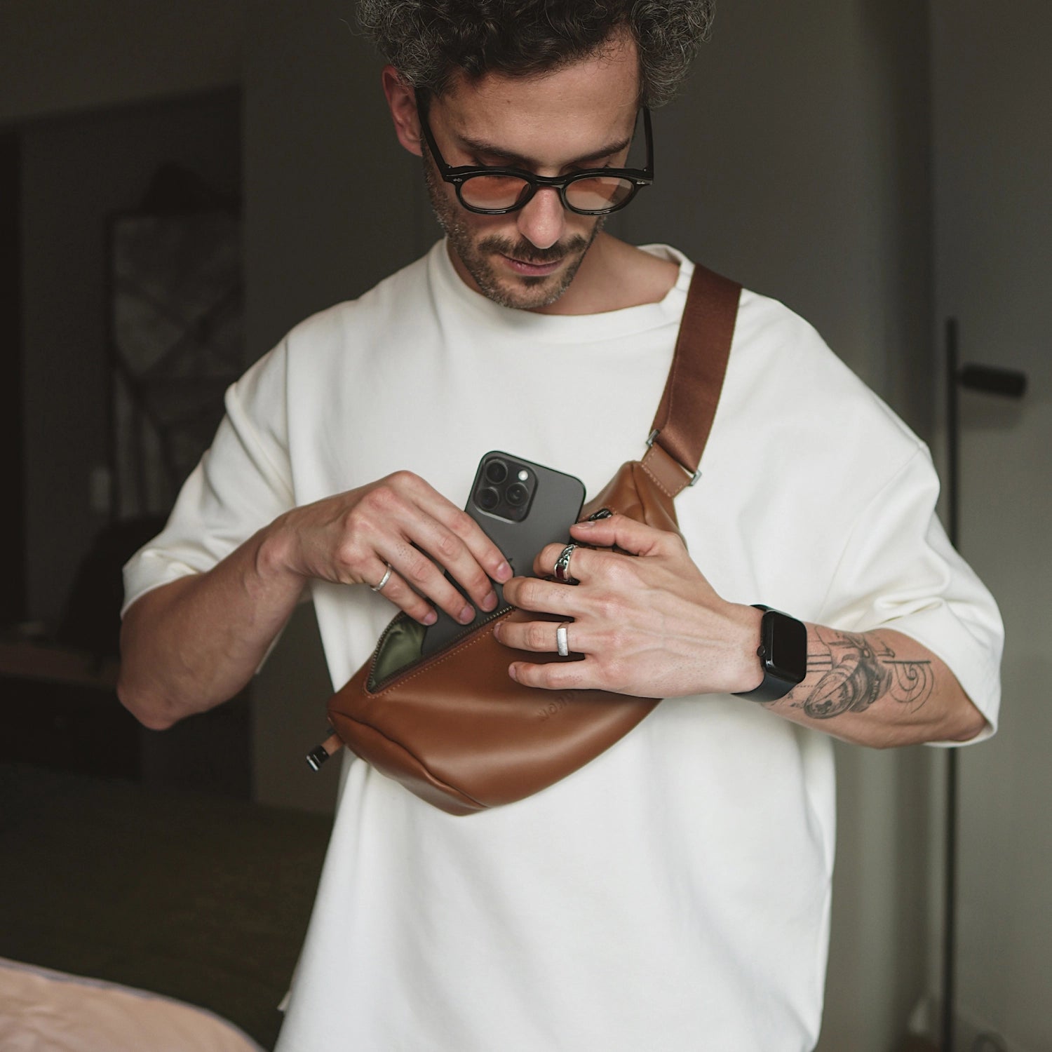 Man inserting an iPhone into the Nordweg Pulse Leather Sling Bag in caramel while wearing it crossbody